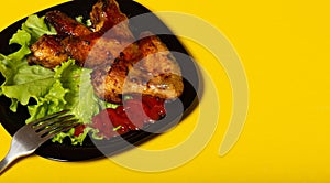 fried chicken wings with salad and ketchup in a black plate and fork on a yellow background. delicious banner