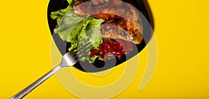 fried chicken wings with salad and ketchup in a black plate and fork on a yellow background. delicious banner