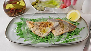 Fried dorado fillet with crispy skin. Woman pouring olive oil on a fish. Arugula, cherry tomatoes, olives, lemon and olive oil.