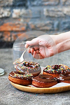 Fried doughnuts with chocolate glaze. Woman`s hand decorating doughnuts with colorful sprinkles