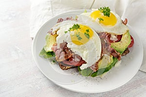 Fried egg, bacon and avocado on a dark whole meal bread with lettuce and tomato, delicious breakfast sandwich, light background,