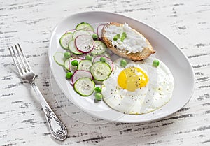 Fried egg, salad with cucumbers, radishes and green peas, toast with feta cheese on a light ceramic plate on wooden background.