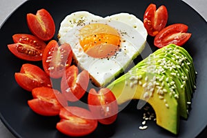 Fried egg in the shape of a heart next to sliced avocado and cherry tomatoes on a black plate