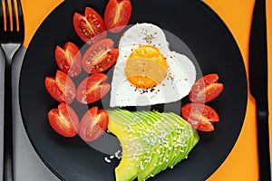 Fried egg in the shape of a heart next to sliced avocado and cherry tomatoes on a black plate