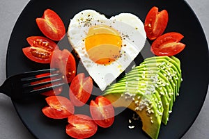 Fried egg in the shape of a heart next to sliced avocado and cherry tomatoes on a black plate