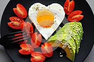 Fried egg in the shape of a heart next to sliced avocado and cherry tomatoes on a black plate