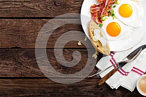 Fried eggs, bacon and italian ciabatta bread on white plate. Cup of coffee. Breakfast. Top view. Wooden background