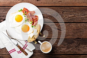 Fried eggs, bacon and italian ciabatta bread on white plate. Cup of coffee. Breakfast. Top view. Wooden background