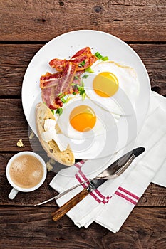 Fried eggs, bacon and italian ciabatta bread on white plate. Cup of coffee. Breakfast. Top view. Wooden background