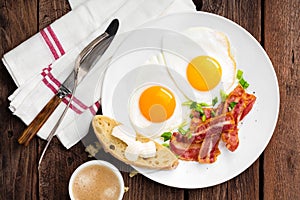 Fried eggs, bacon and italian ciabatta bread on white plate. Cup of coffee. Breakfast. Top view. Wooden background
