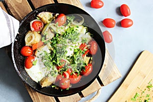 Fried eggs with cherry tomatoes, potatoes and onions in a pan on a wooden stand on the table