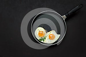 Fried eggs in frying pan with arugula leaves, salt and pepper for breakfast on black wooden background. Top view. Flat lay. Copy