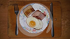 Fried eggs from one egg with pieces of fried bacon on a plate with a knife and fork on a wooden cutting board