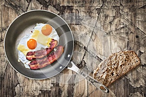 Fried Eggs and Pork Bacon Rashers in Teflon Frying Pan with slice of Integral Bread on Old Cracked Peeled Wooden Table photo