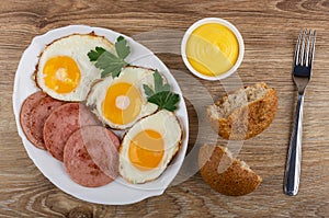 Fried eggs with sausages, parsley in dish, bowl with mayonnaise, broken bun, fork on table. Top view