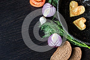 Fried eggs in the shape of a heart in a frying pan, near which lay bread slices, red onion, tomato, garlic and dill