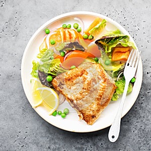 Fried fillet of sea white cod fish with juicy lettuce, capsicum, lemon, green peas on a large white dish on a gray