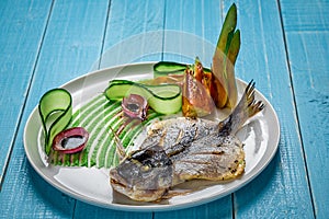 Fried fish dorado, decorated with cucumbers and onions. On a blue wooden background