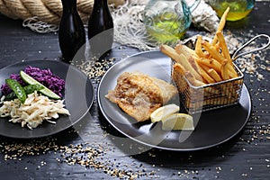 Fried fish fillet served with golden fries in a metal serving basket and salad mix, on black plates, selective focus.