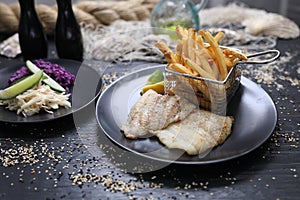 Fried fish fillets served with potato fries in a metal serving basket and salad mix, on black plates, selective focus.