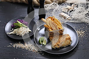 Fried fish fillets served with potato fries in a metal serving basket and salad mix, on black plates, selective focus.