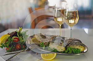 Fried fish on metallic dish with greens and lemon on white table. Indoors.