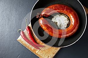 fried meat sausage in a cast iron pan and hot chili pepper on a cutting wooden board on a dark stone table