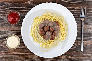 Fried meatballs with spaghetti in dish, fork, bowls of ketchup and mayonnaise on wooden table. Top view