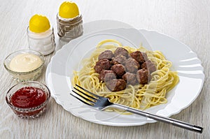 Fried meatballs with spaghetti, fork in dish, salt, pepper, bowls with ketchup and mayonnaise on wooden table