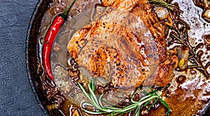 Fried pork meat in frying pan over dark background, top view. Chili pepper and rosemary