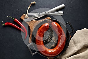 Fried pork sausage tied with a ring on a black cast iron pan on a wooden cutting board with hot peppers and cutlery on a