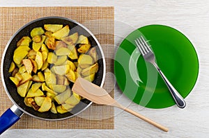 Fried potatoes in frying pan, spatula on mat, plate and fork on wooden table. Top view