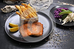 Fried rainbow trout fillet served with potato fries in a metal serving basket and salad, on black plates. Fish and chips
