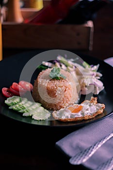 Fried rice, fried egg, cucumber, sliced tomato, green salad and Fried chicken placed on a black plate as a garnish