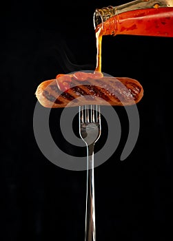 Fried sausage on a fork. Isolated on a black background. Cholesterol, junk food. Dynamics in the frame, pouring hot sauce ketchup