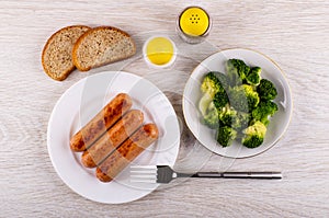 Fried sausages in plate, fork, broccoli in plate, bread, salt, pepper on table. Top view