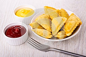 Fried savory pies in plate, bowls with mayonnaise and ketchup, fork on table