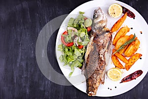 Fried sea bass with lemon, potatoes, salad and spices on a black wooden table