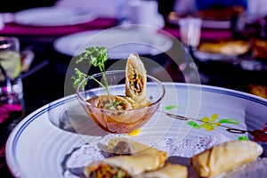 Fried spring rolls with red and white sauces, served in white plate with fresh green salad over gray background. Asian