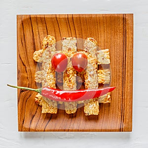 Fried tempeh decorated with chilly and tomato smile on a wooden tray. Top view. Close-up. Square image.