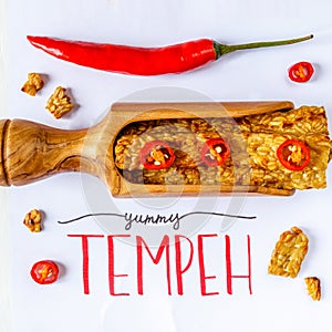 Fried tempeh in a wooden scoop decorated with chilly. YUMMY TEMPEH caption. Top view.