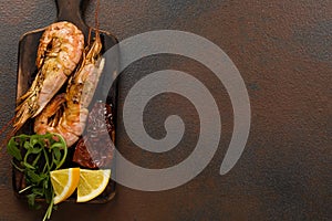 Fried tiger shrimps with lemon, sun-dried tomatoes and greens or rukkola on a dark wooden cutting board.