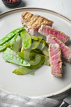 Fried tuna steak in sesame with spring onions and sugar snap peas, on plate, on white stone  background