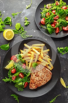 Fried Tuna Steaks on Black Plate with Fresh Green, Tomato Salad, lemon and french fries. healthy sea food