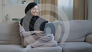 Friendly arabian muslim woman girl in hijab sit on cozy sofa talk video call with friend greeting at computer webcam