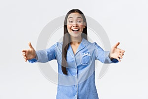 Friendly asian girl in blue pajamas welcome girlfriends to her sleepover party, smiling happy and reaching hands forward