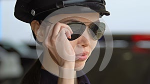 Friendly asian policewoman in hat putting on sunglasses and smiling, trust