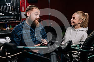 Friendly bearded cycling repairman male having conversation with positive blonde female client, talking about problem of