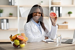 Friendly black female dietitian holding grapefruit and smiling at camera, offering online weight loss consultations