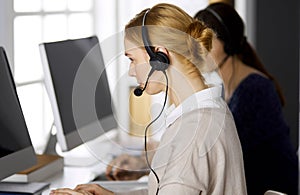 Friendly businesswoman talking by headset while sitting in office. Call center and diverse people group in business